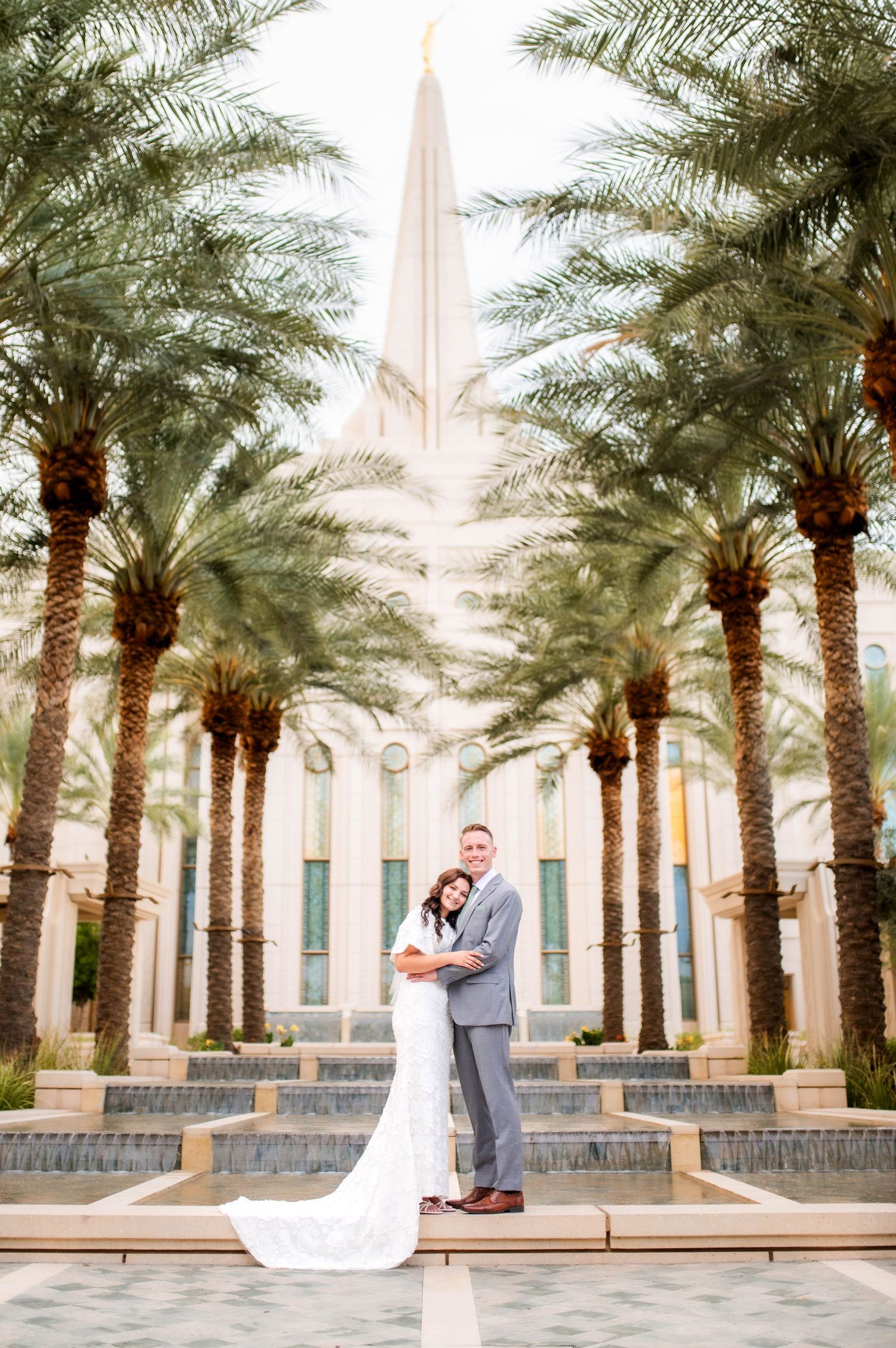 Bride and groom pose in front of an LDS temple surrounded by tall palm trees on their wedding day.