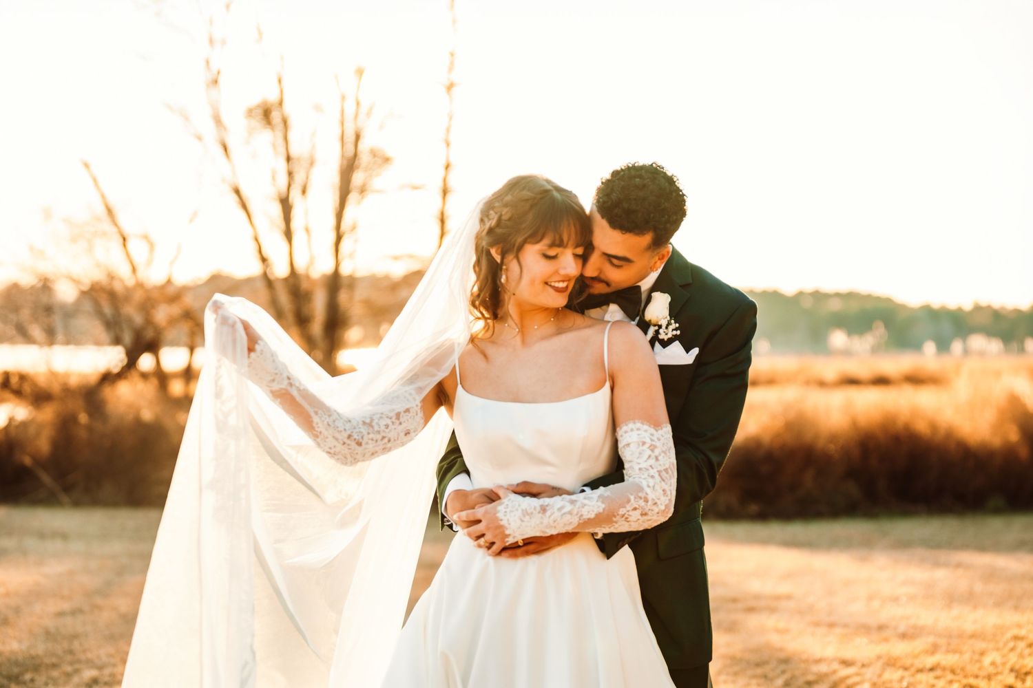 Bride and groom share a romantic golden hour embrace outdoors, her lace veil flowing in the breeze.