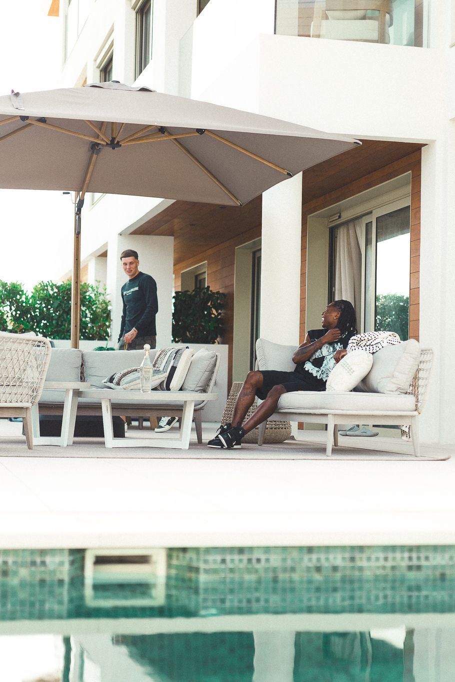 Two people relaxing on a modern outdoor patio with pool, white furniture, and large umbrella at a luxury villa.