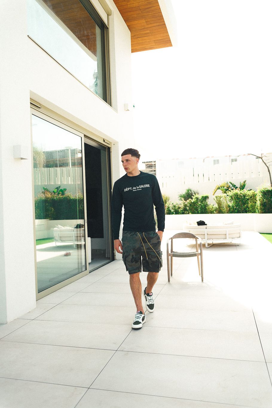 Young man in black sweatshirt and camo shorts walking on modern patio with large glass doors and outdoor furniture.