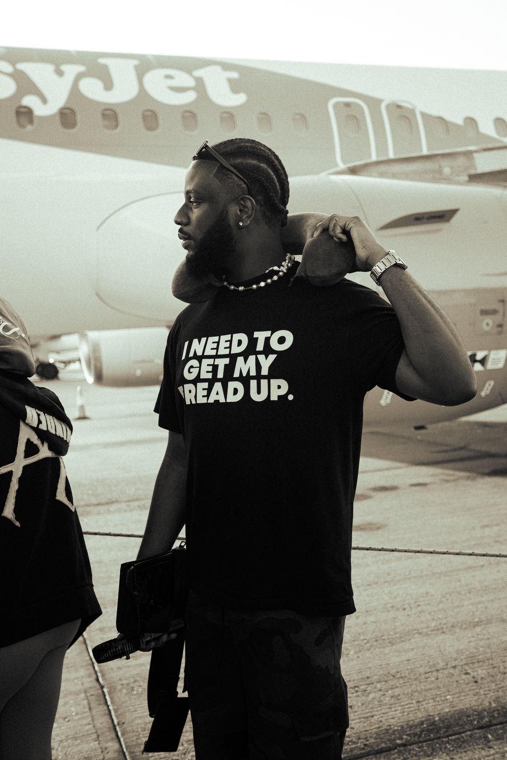 Man wearing black t-shirt reading I Need To Get My Bread Up stands on airport tarmac near easyJet plane.