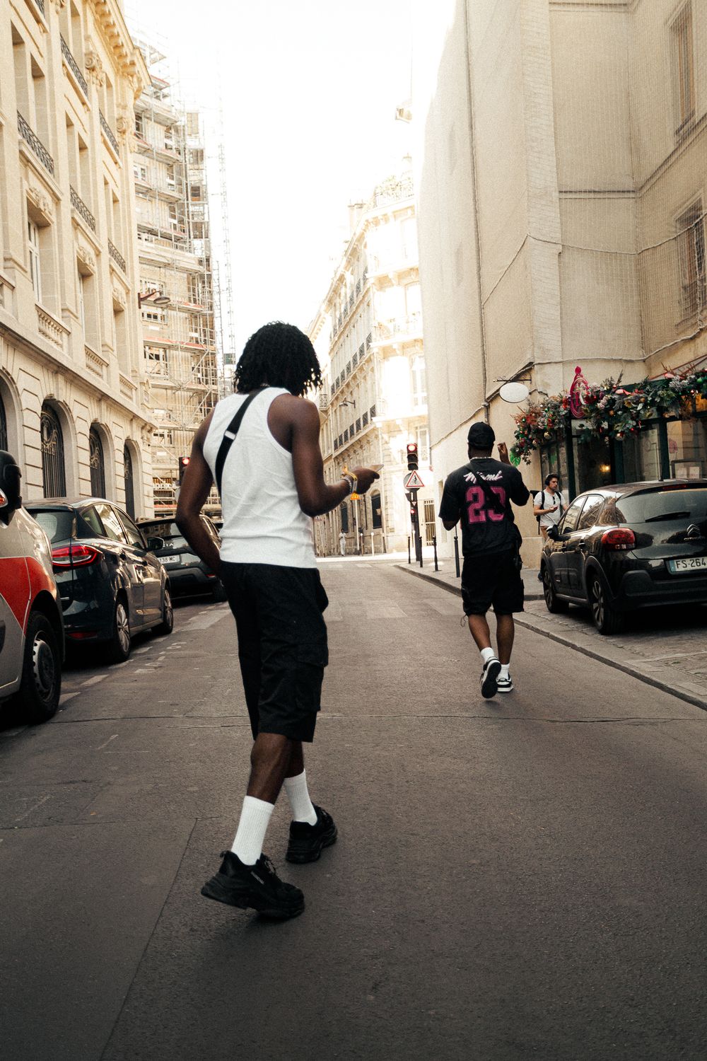 Two men walking down a narrow Parisian street lined with parked cars and classic Haussmann buildings on a sunny day.