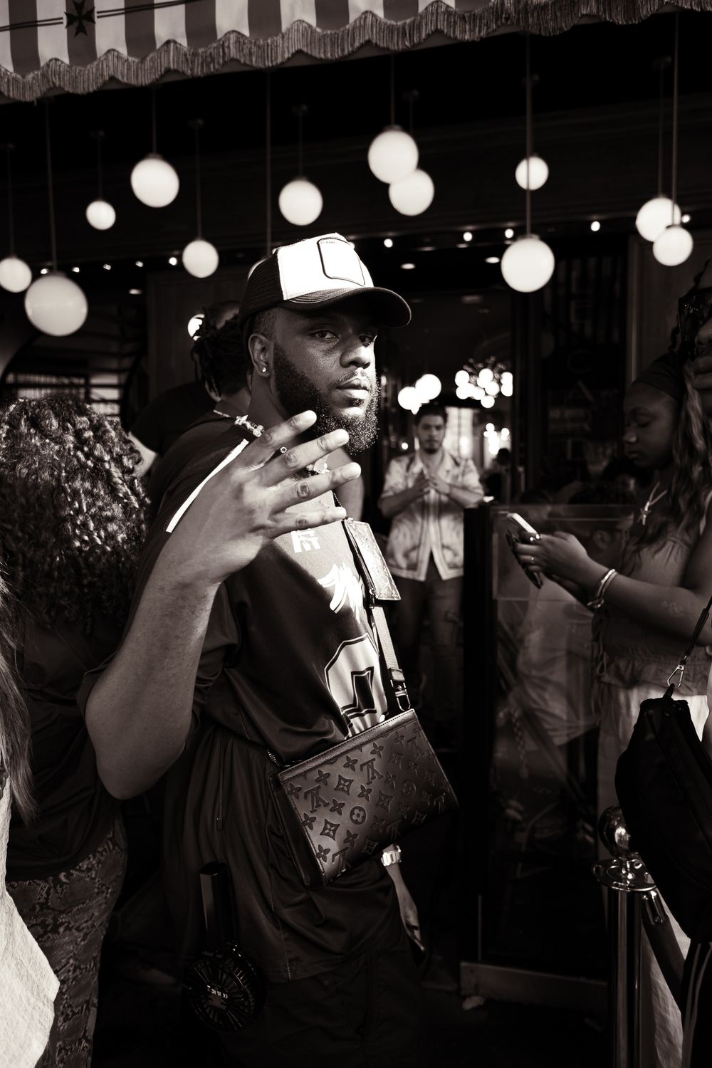 Black and white photo of a bearded man wearing a cap and Louis Vuitton bag, posing with hand gesture at a nightclub.