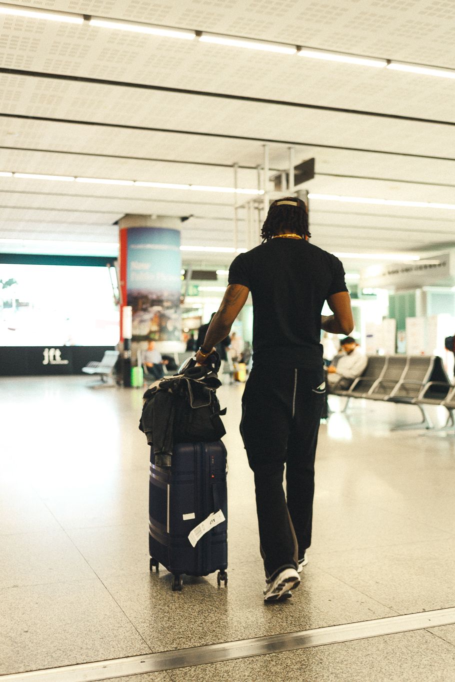 A traveler dressed in black walks through an airport terminal pulling a blue suitcase with a bag on top.