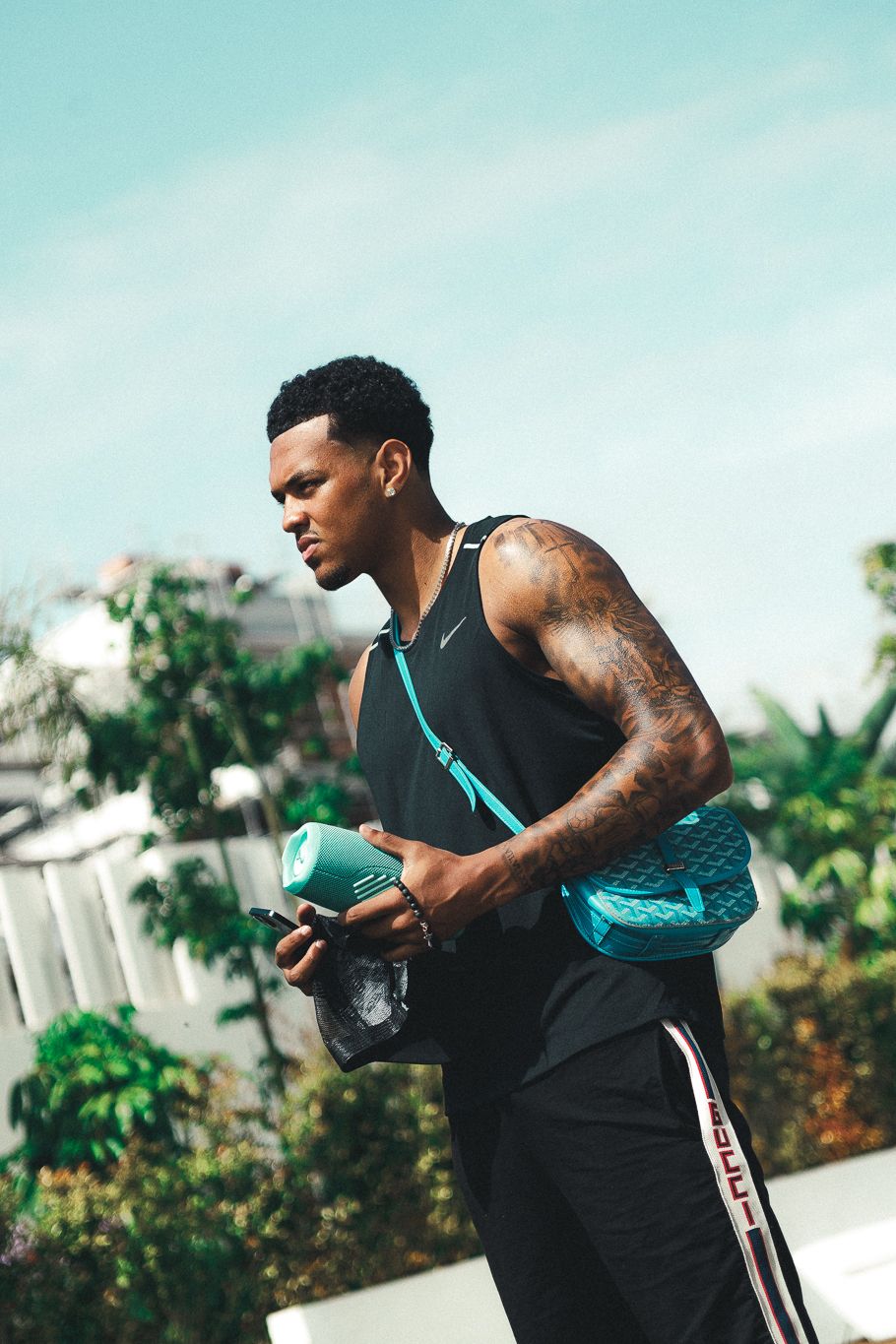 Tattooed muscular Black man in black tank top holds gym bag and phone outdoors on sunny day.