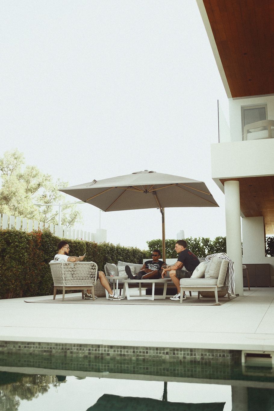 Three people relaxing on modern outdoor furniture under a large umbrella beside a pool at a luxury home.