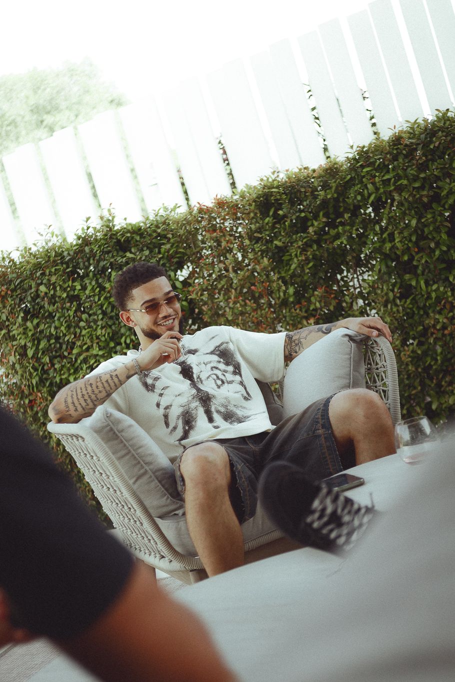 Young man with tattoos and sunglasses smiling while relaxing in a white wicker chair outdoors near a green hedge.