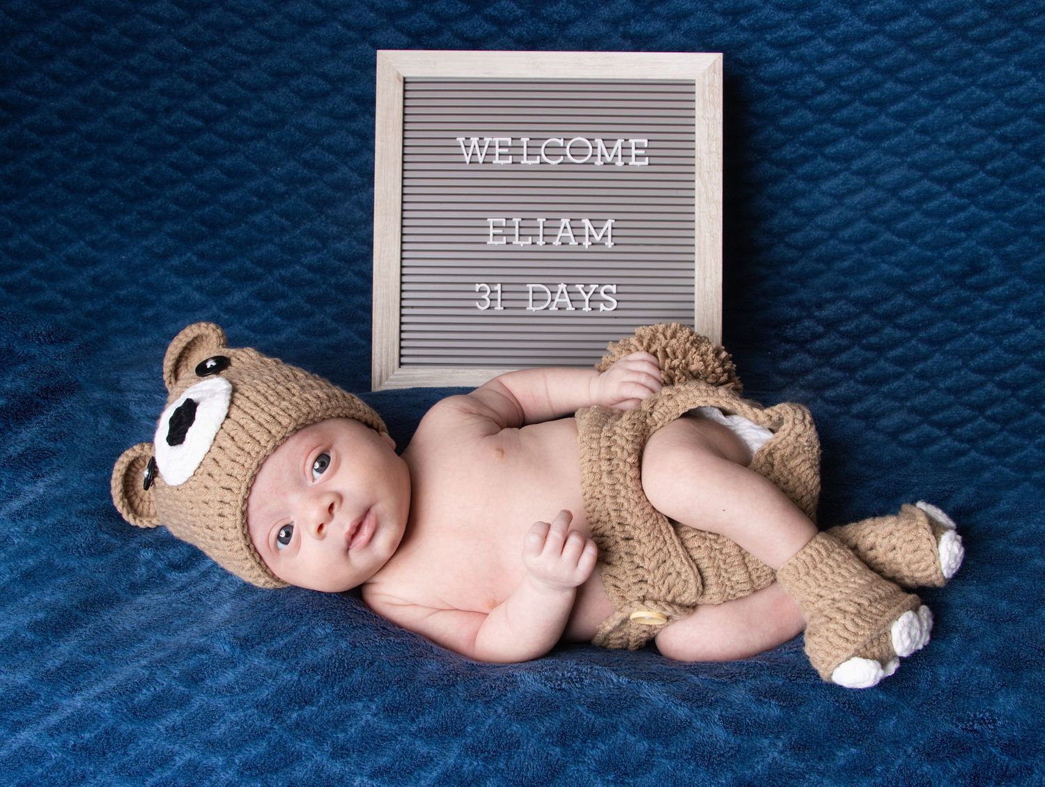 Newborn baby Eliam at 31 days old wearing a knit bear outfit posed next to a welcome letter board.