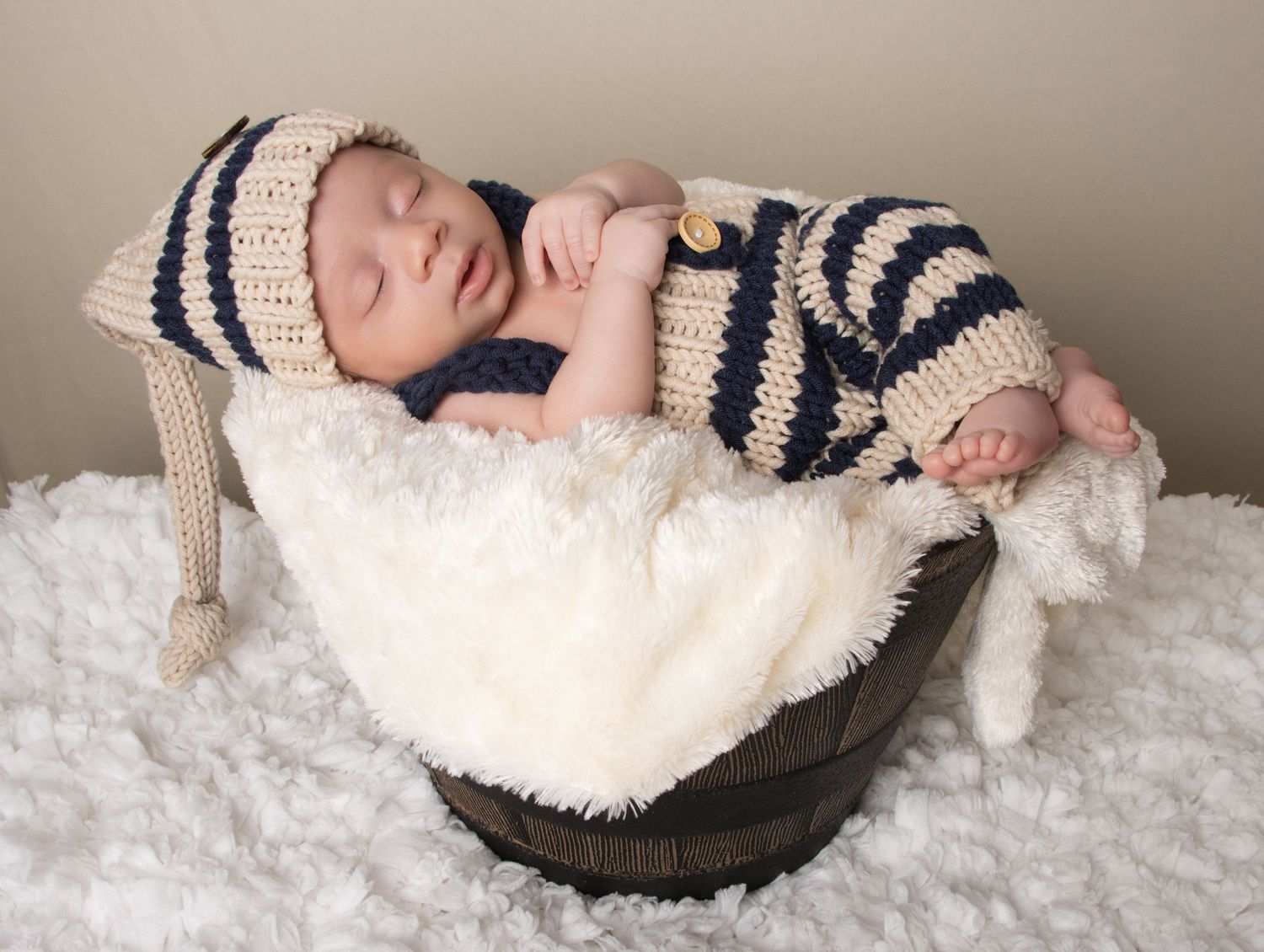 Sleeping newborn baby in striped knit outfit and hat, nestled in a wooden bucket with white fluffy blanket.