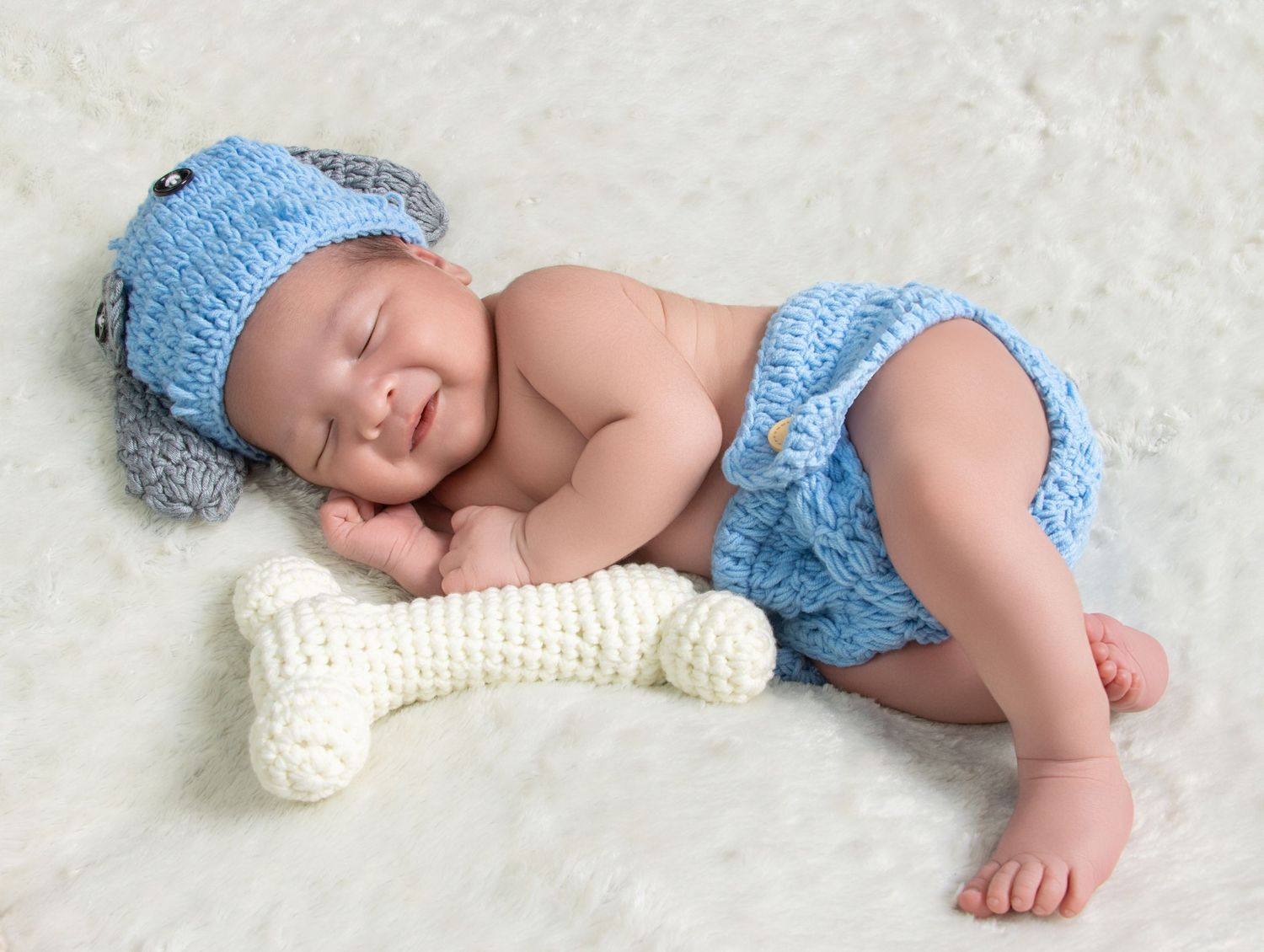 Smiling newborn baby wearing blue knit puppy hat and diaper cover, sleeping with a crocheted bone prop.