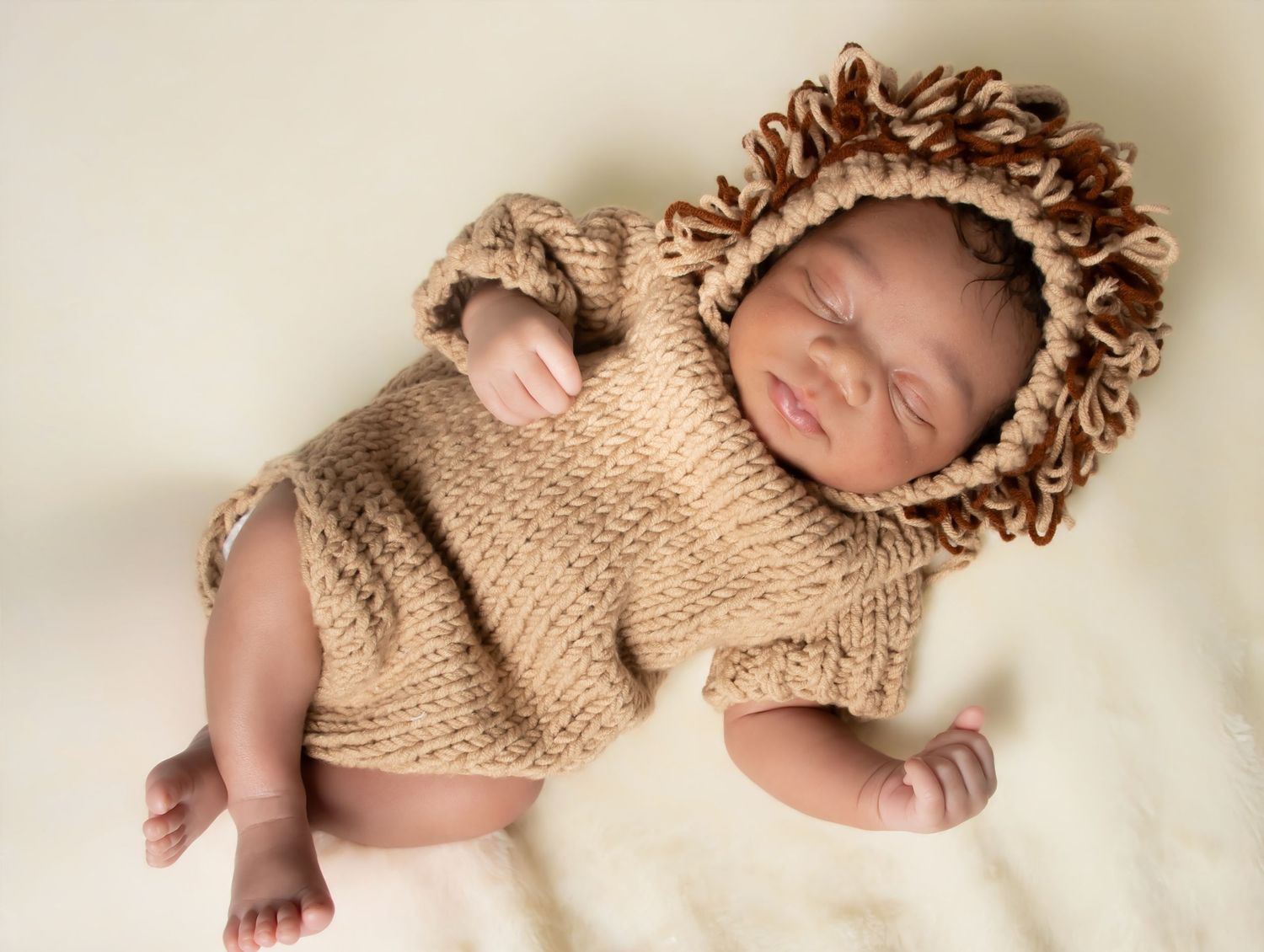 Newborn baby sleeping peacefully in a knitted lion costume with mane hat on a white fluffy blanket.