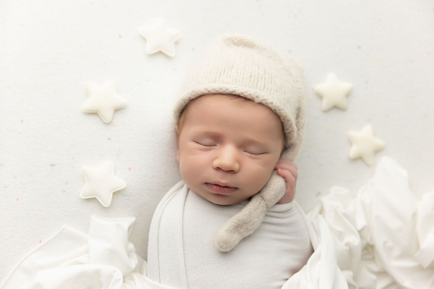 Sleeping newborn baby in white knit hat surrounded by delicate white paper stars on soft white blanket.