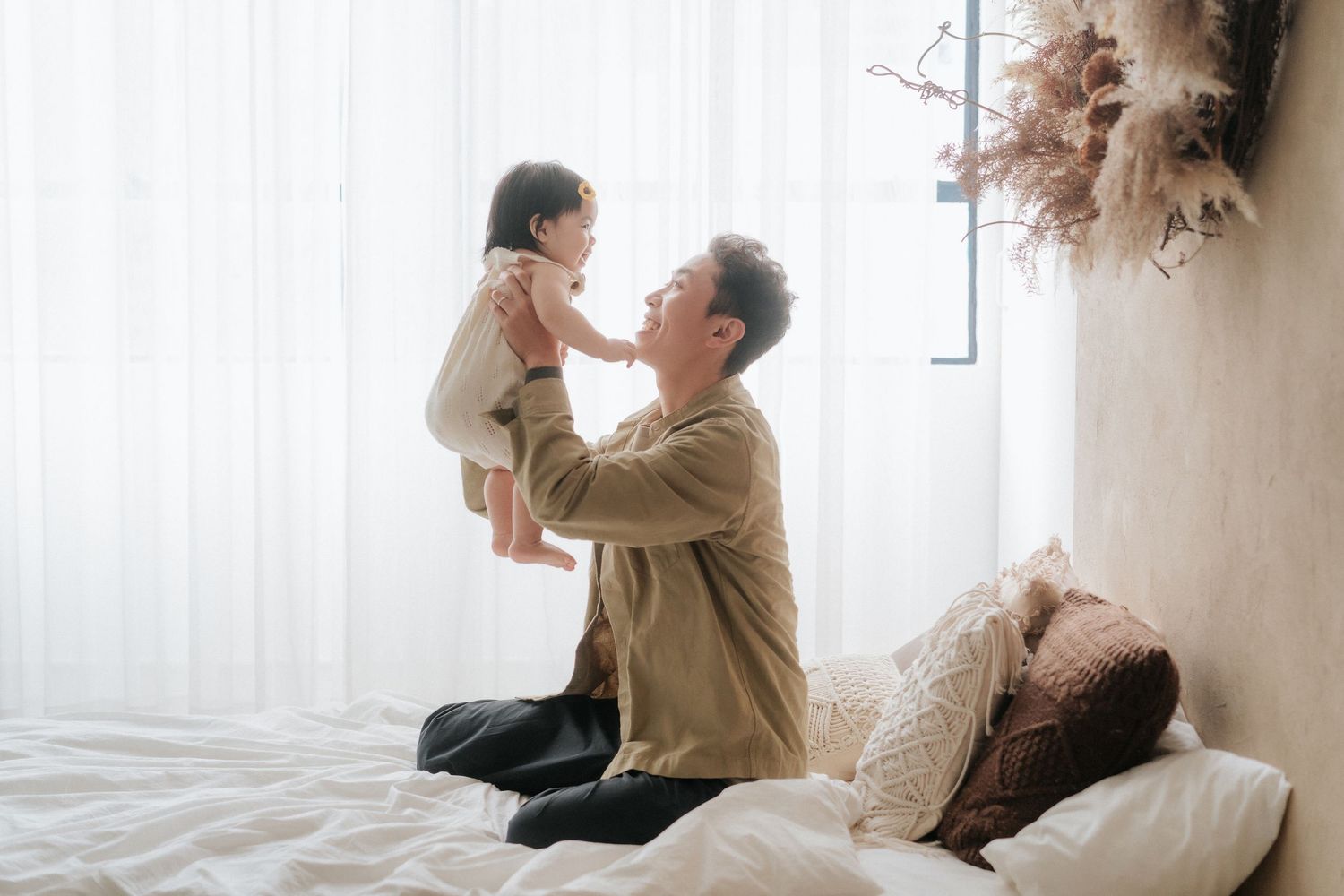 A parent and child share a tender moment while playing together on a bed near a window with sheer curtains.