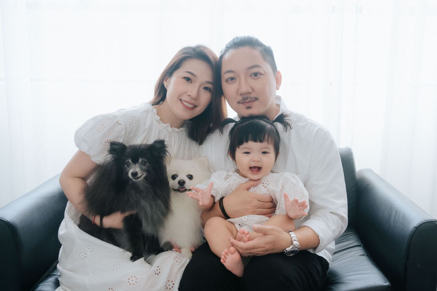 Happy Asian family with baby and black Pomeranian dog posing together on sofa in bright white studio.幸福的亞洲家庭帶著寶寶和黑色博美犬在明亮的白色攝影棚裡一起坐在沙發上合照。