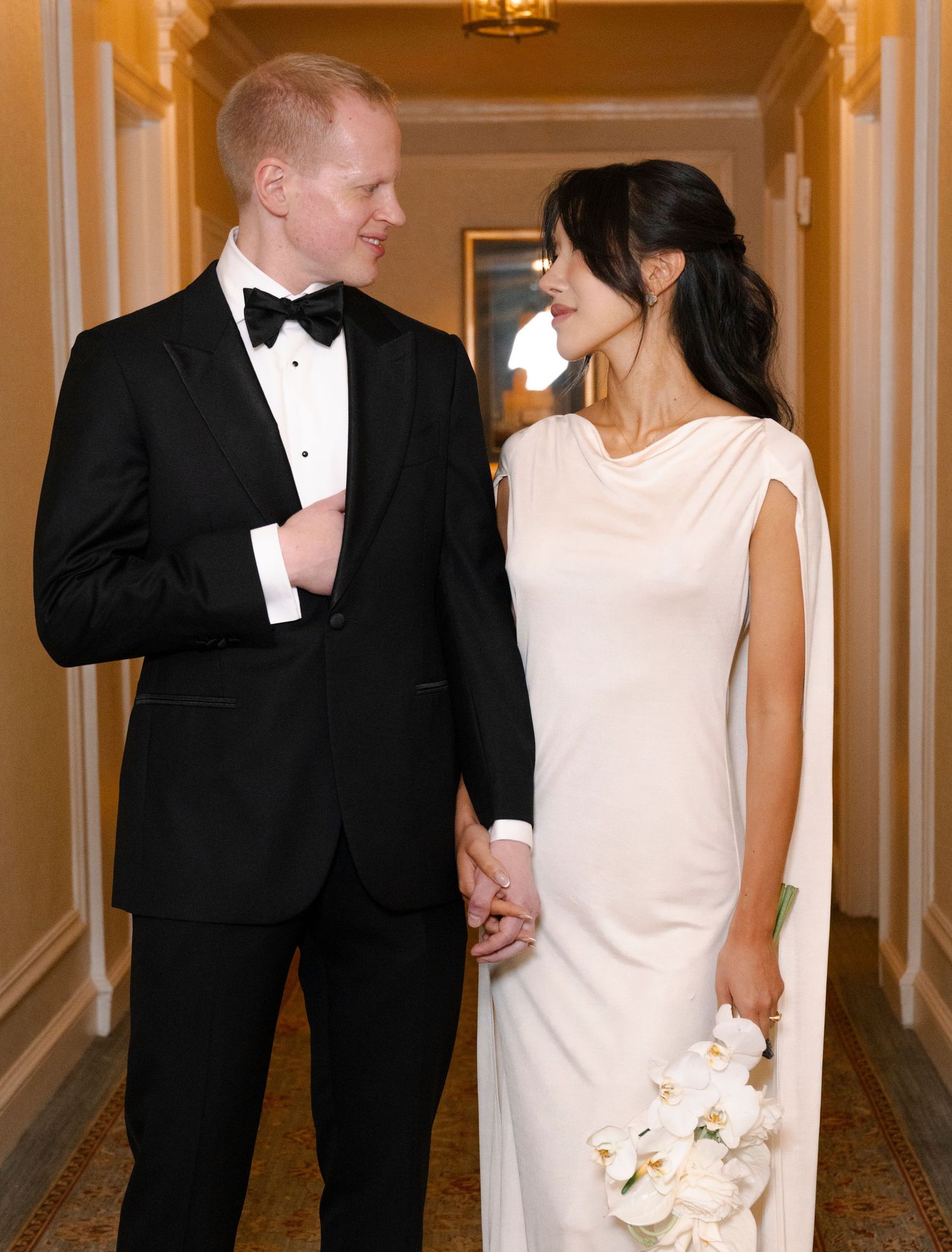 A couple in formal attire smiling at each other in an elegant hallway, with the bride holding a white bouquet.