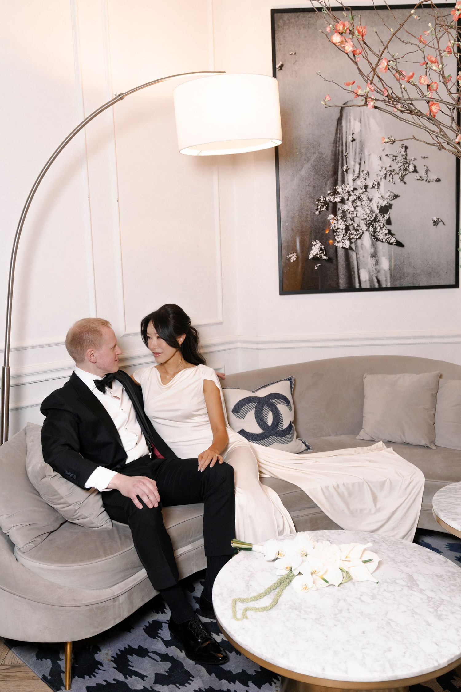 Elegant couple in black and white attire sitting on a modern gray couch under an arched floor lamp with artwork behind them.