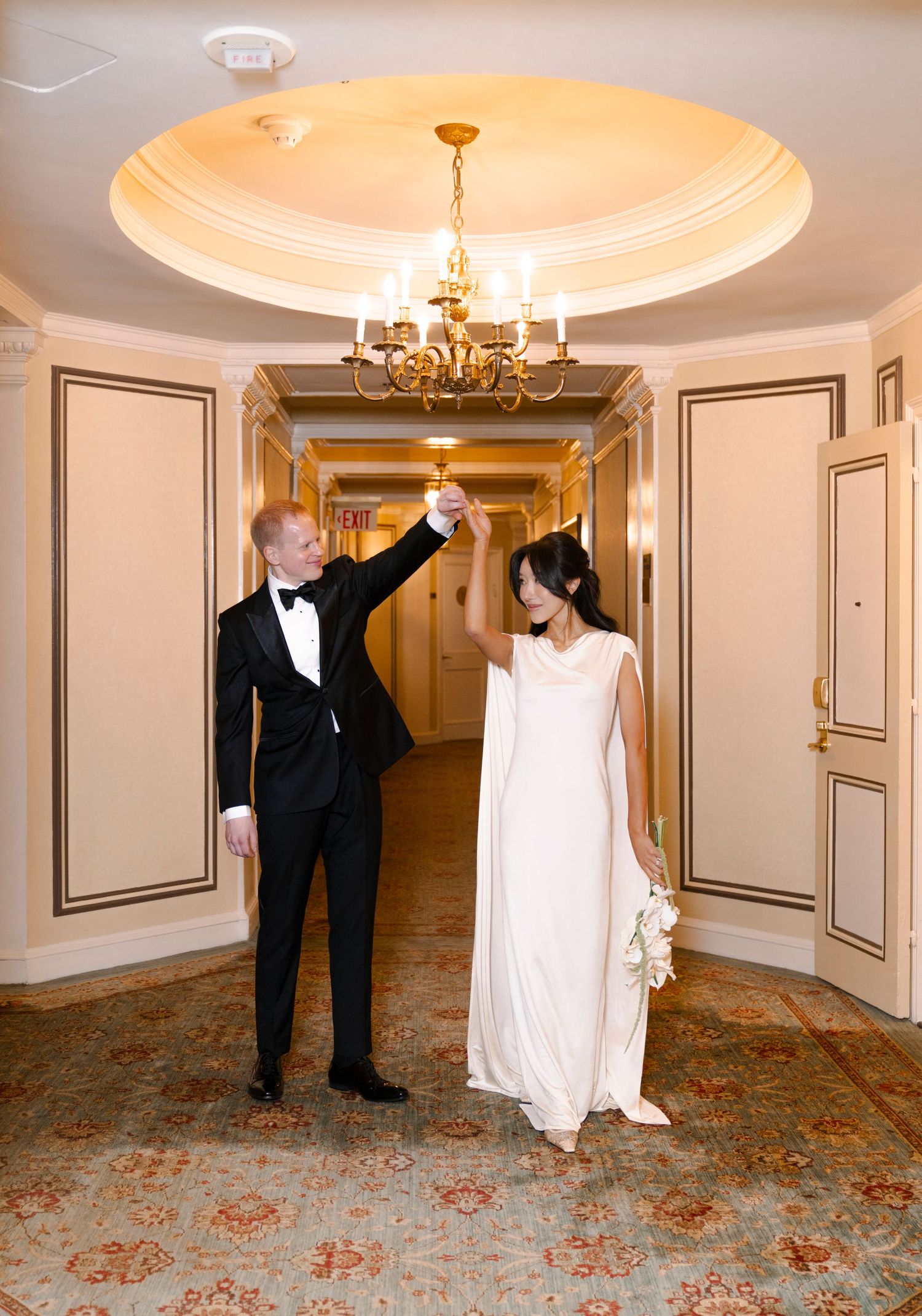 A couple dances together in an elegant hotel hallway with ornate moldings and chandeliers.