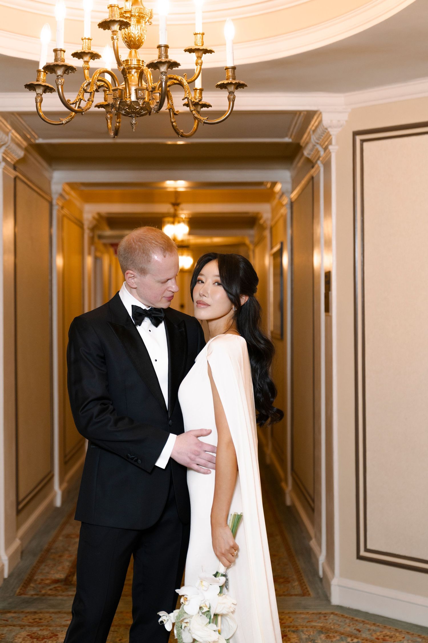 A bride and groom share intimate moments in an elegant hotel hallway with cream walls and chandeliers.