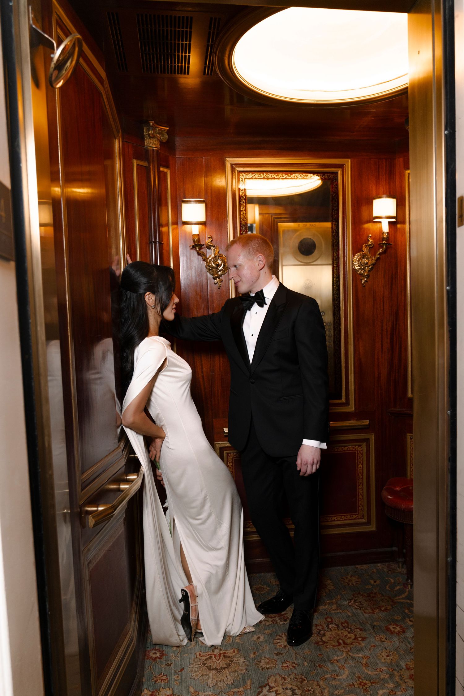 Elegant couple walks through a wood-paneled hallway with ornate lighting fixtures in a formal venue.