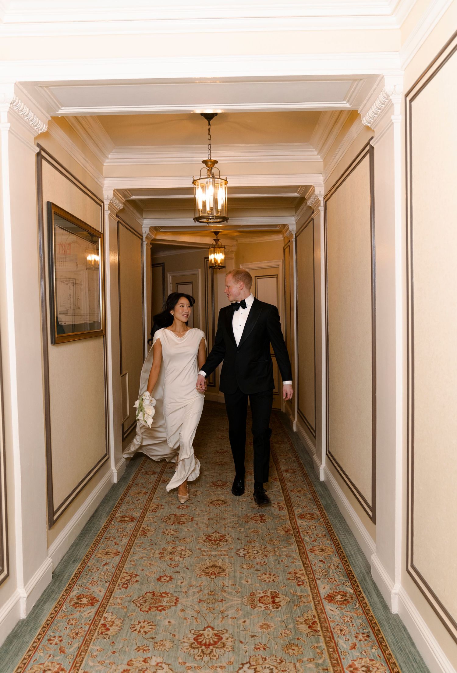 Wedding couple walking down an elegant hotel hallway with cream walls and floral patterned carpet.