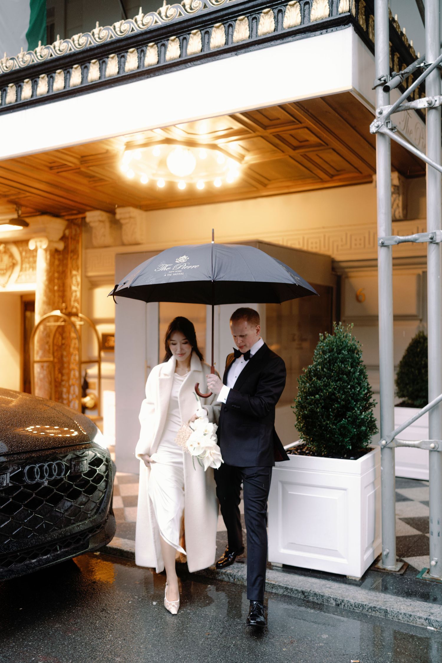 A couple walks under a black umbrella outside an elegant building entrance with ornate architectural details on a rainy day.