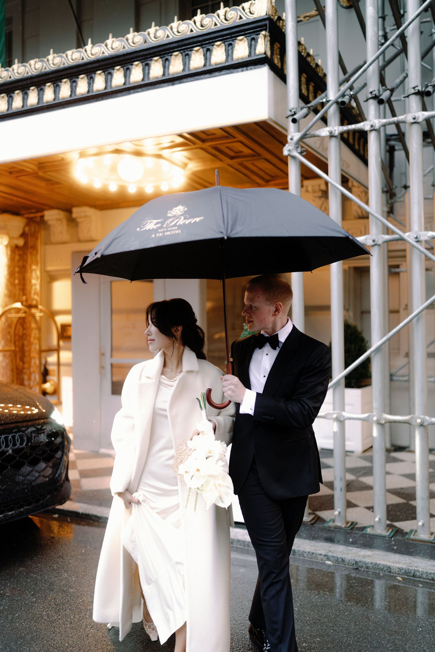 A couple shares a romantic moment under umbrellas in an elegant covered walkway with ornate architectural details.