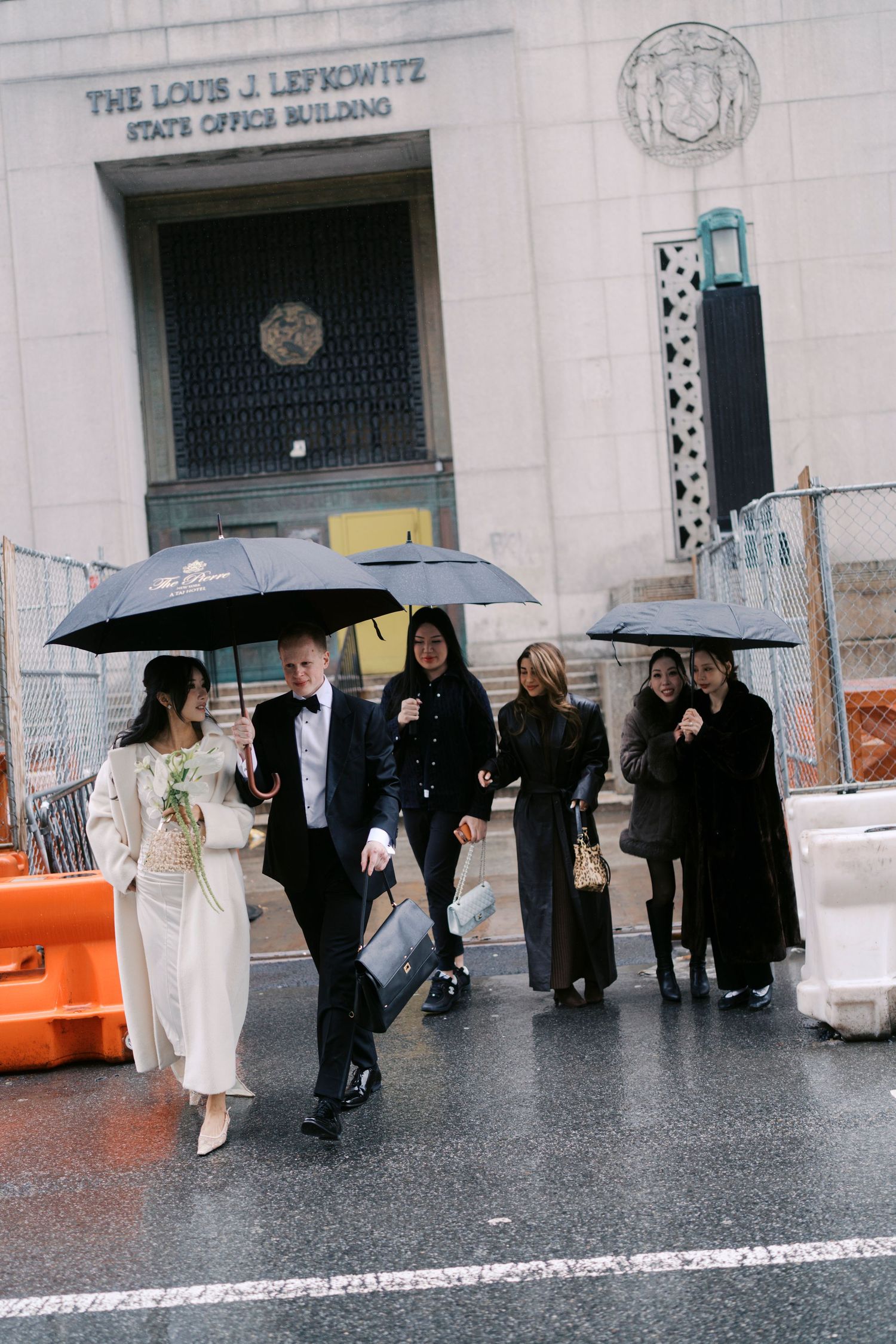 Wedding party with umbrellas gathered outside a New York government building on a rainy day.