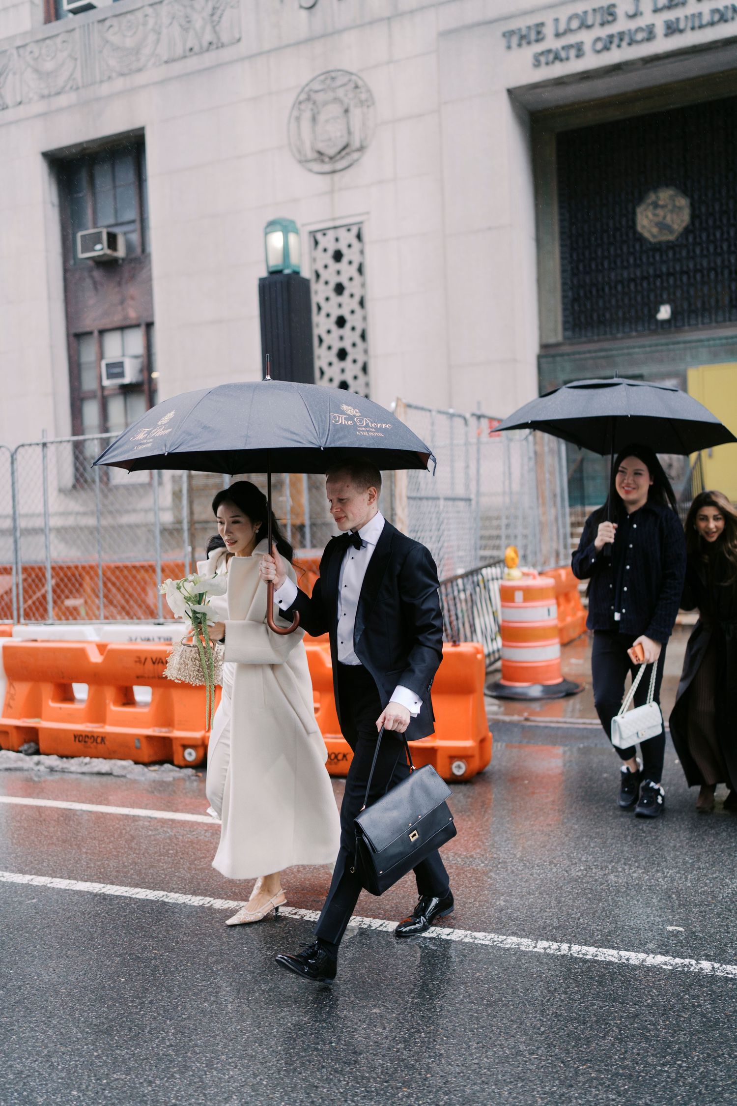 A couple walks with black umbrellas in the rain outside a historic building with orange barriers.