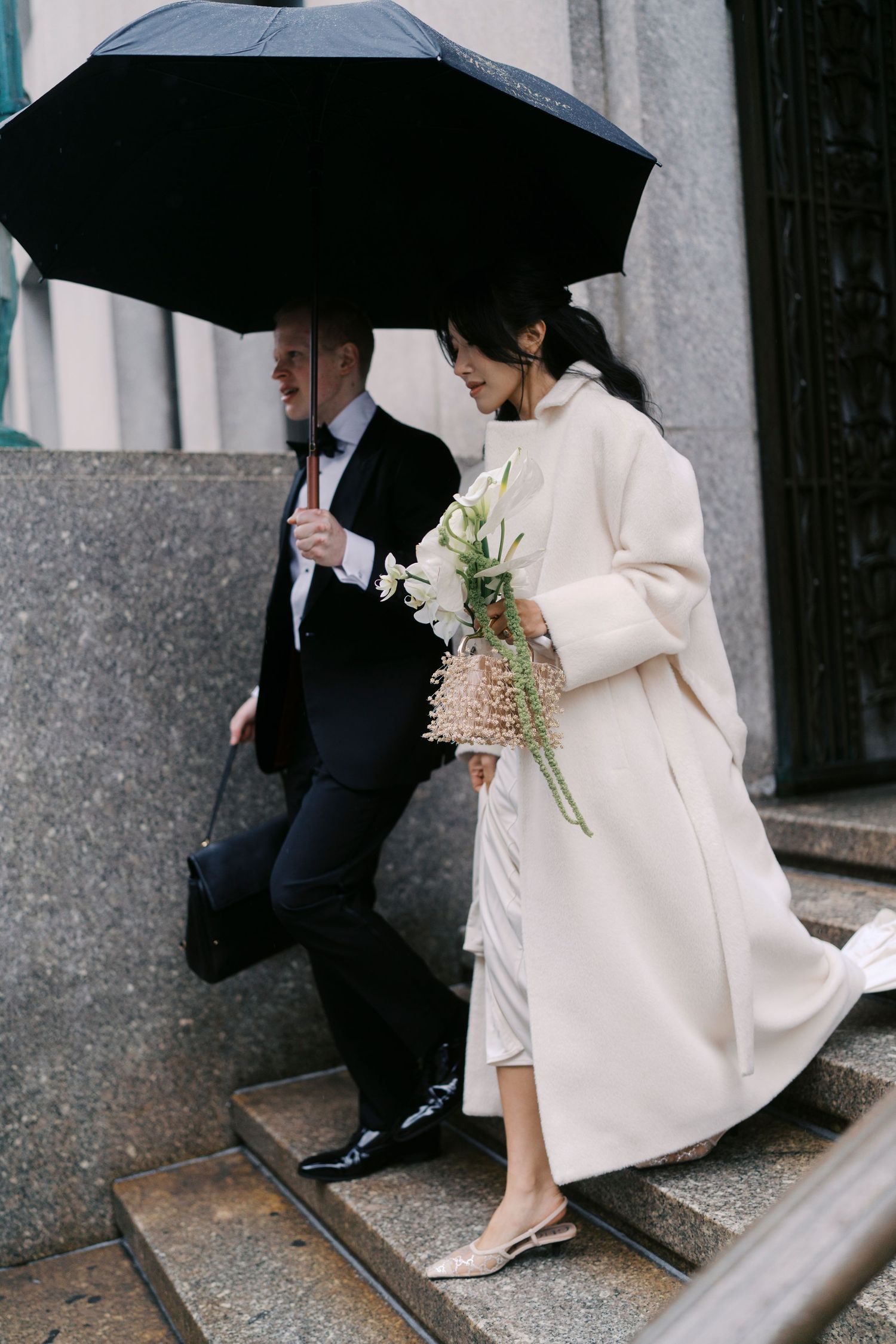 A couple shelters under black umbrellas while walking along a stone building exterior.