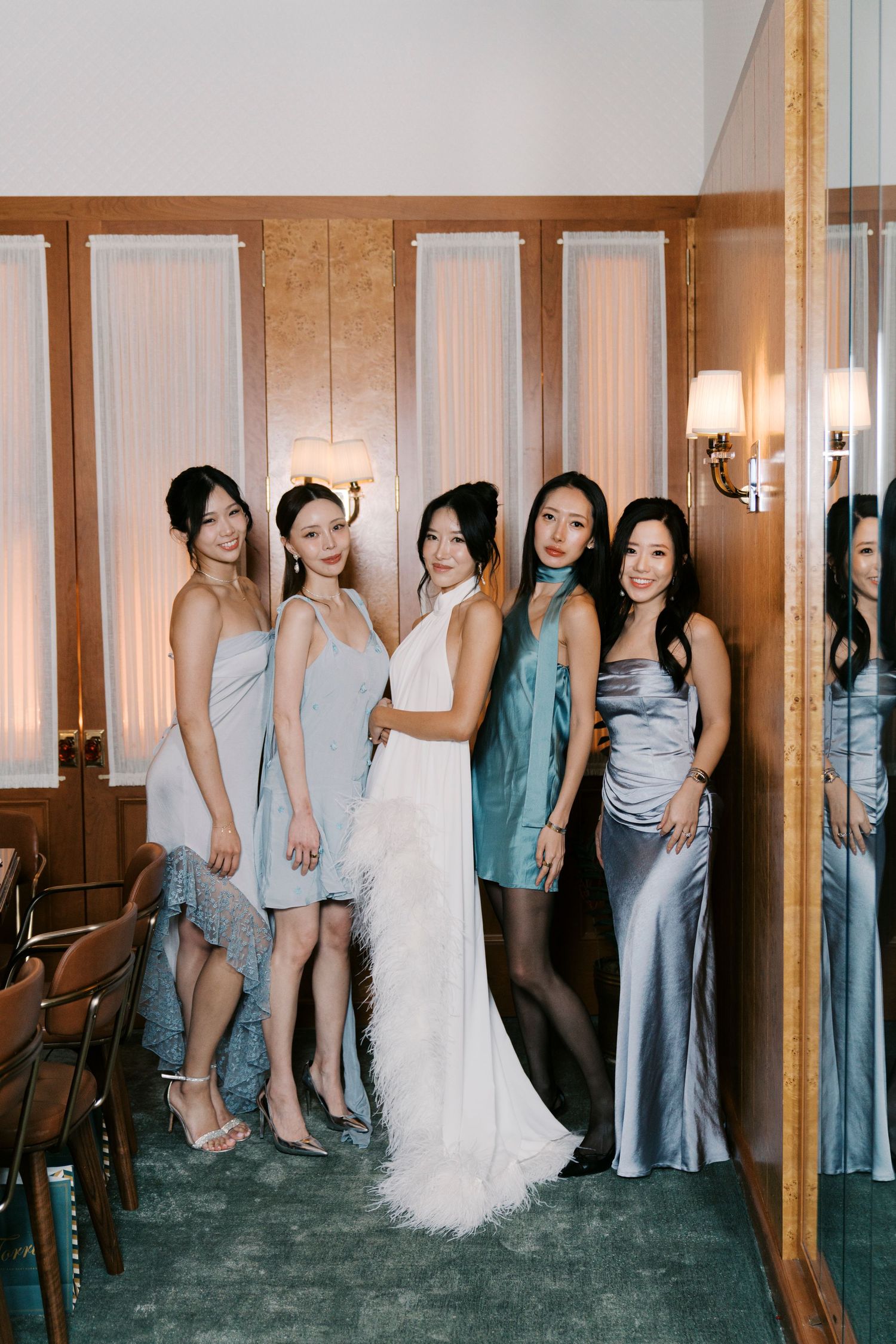 Wedding party group photo showing bridesmaids in blue dresses and white wedding gown against wooden paneled walls.