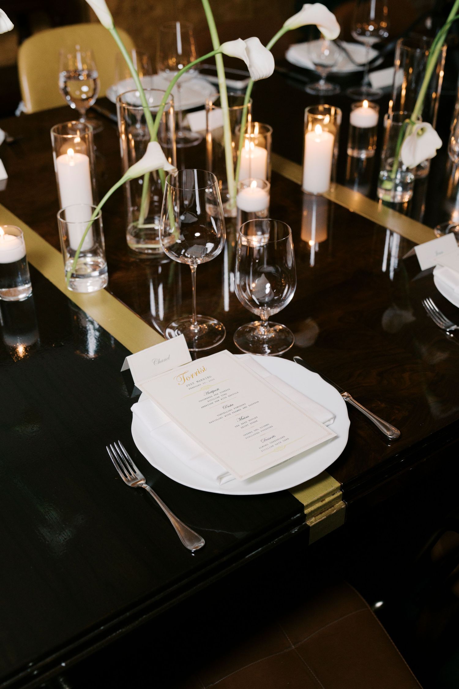 Elegant wedding reception table setting with white menus, glassware, and candlelight on dark tablecloth.