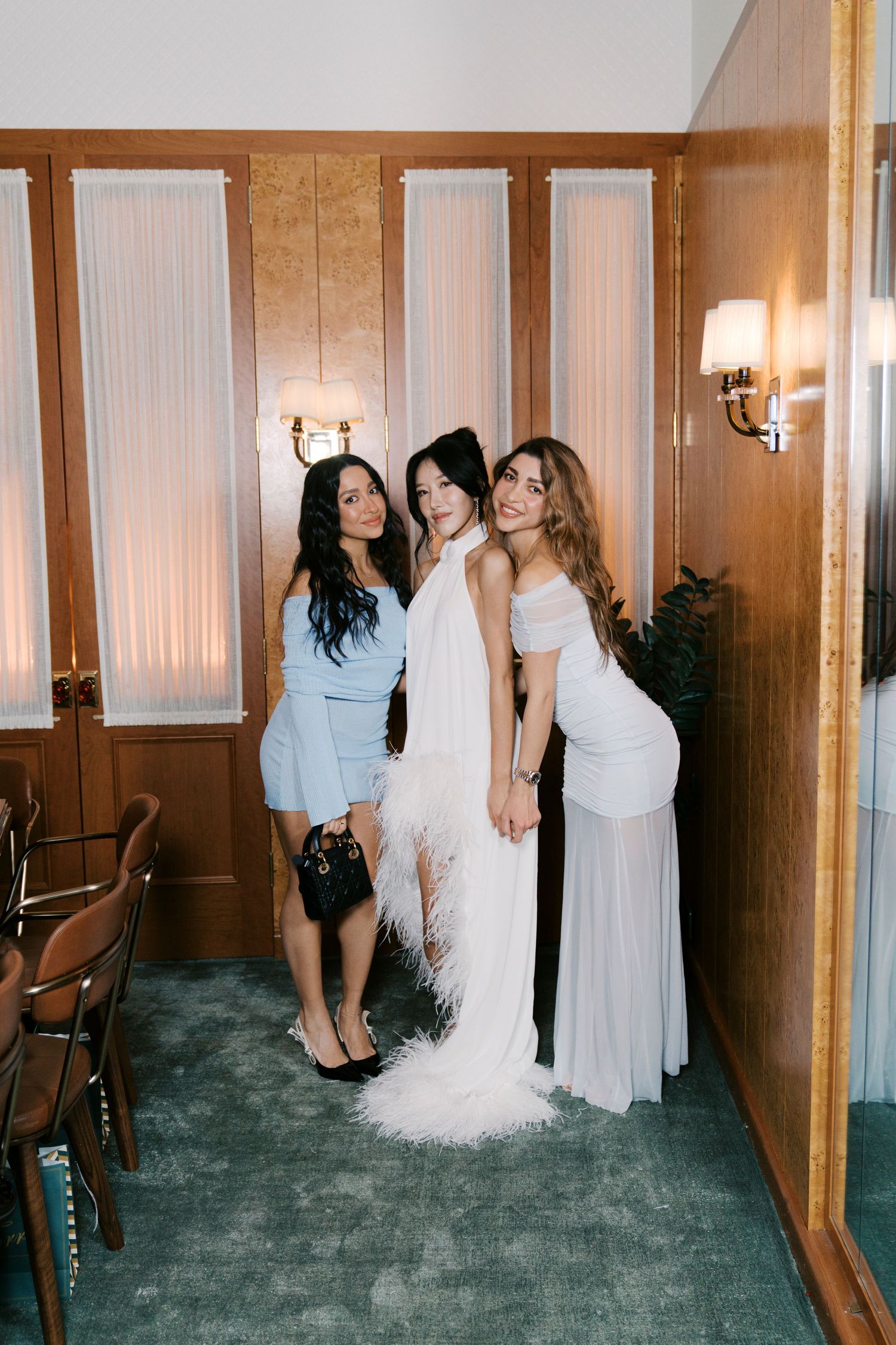 Wedding party members in white dresses gather in a wood-paneled hallway with elegant wall sconces.