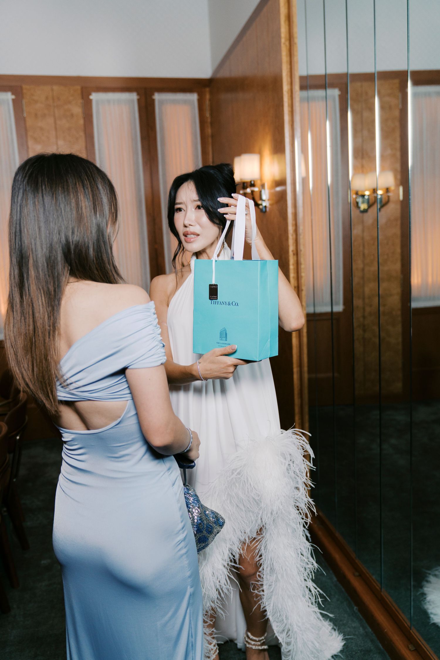 A person in a light blue dress carries a Tiffany shopping bag down a wood-paneled hallway.