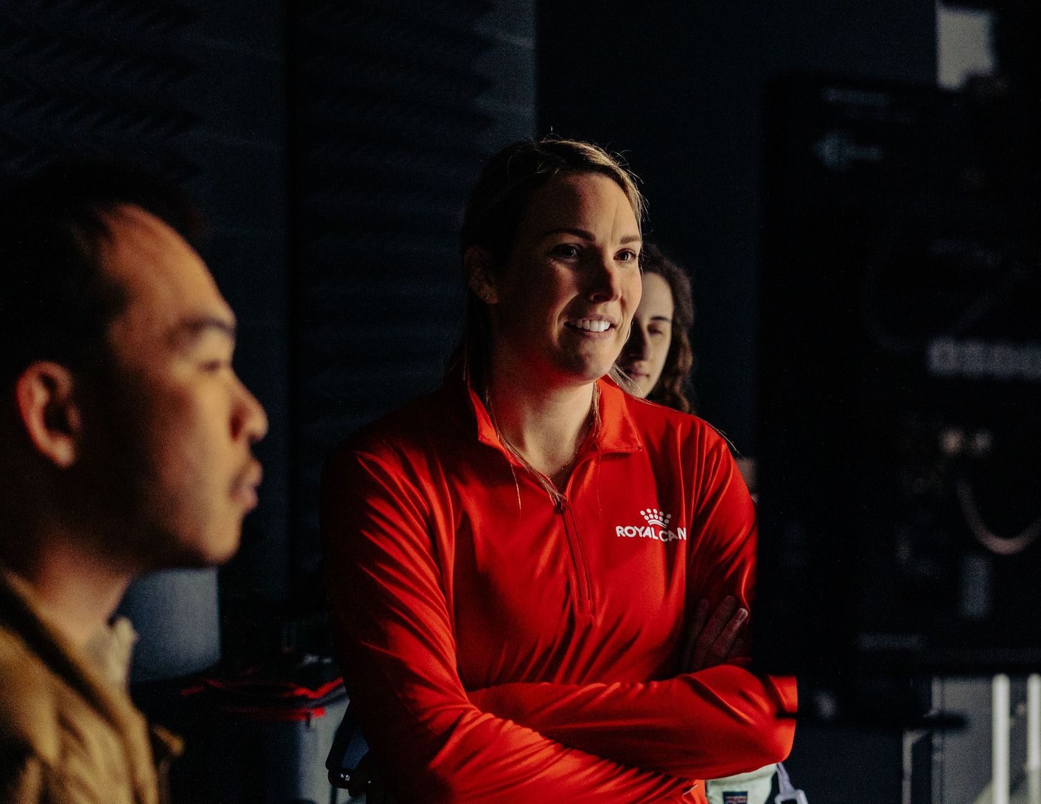 People in a dimly lit room with someone wearing a bright red jacket in focus.