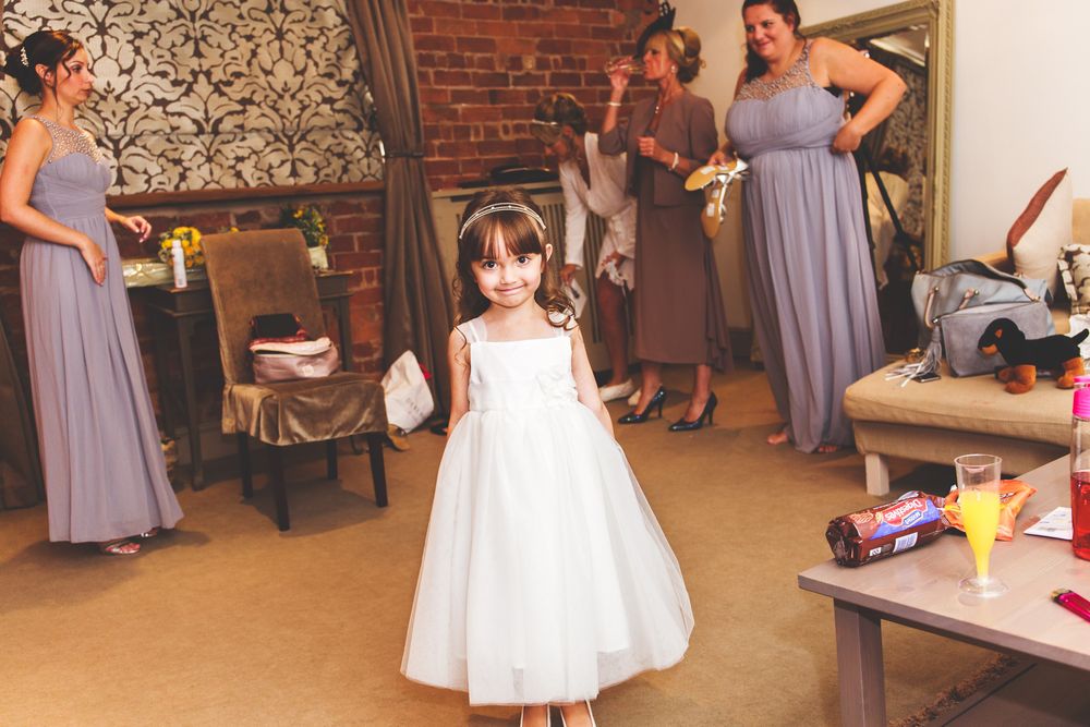 Women in lavender bridesmaid dresses prepare in a rustic brick-walled room.