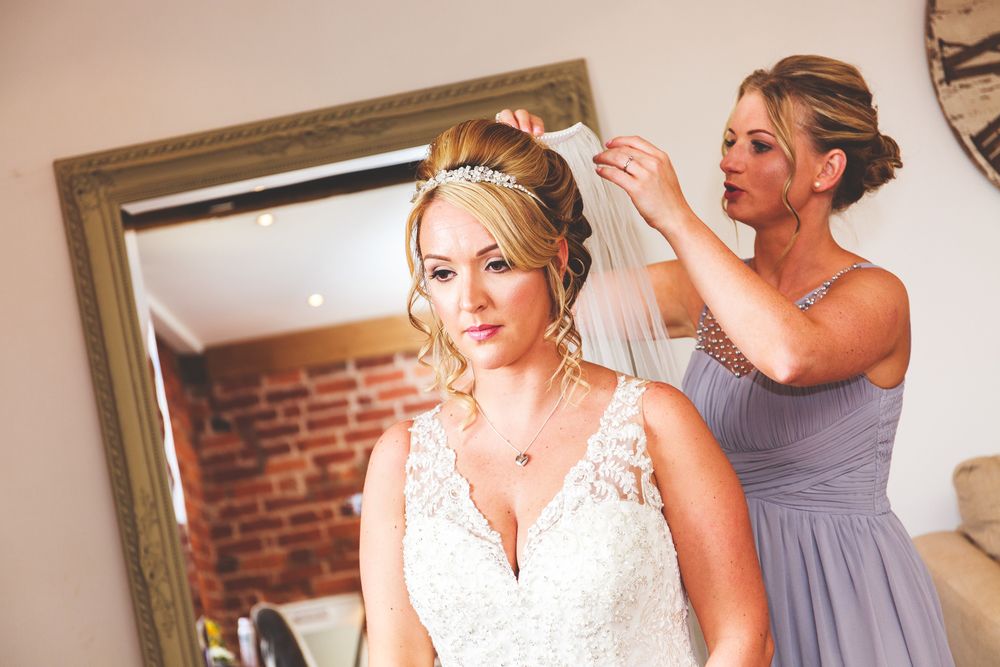 Bride getting ready with help from attendants in a sunlit room with exposed brick walls.
