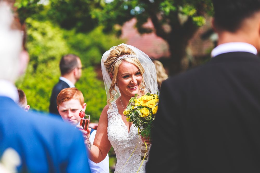 A bride with yellow bouquet waves during an outdoor wedding ceremony.