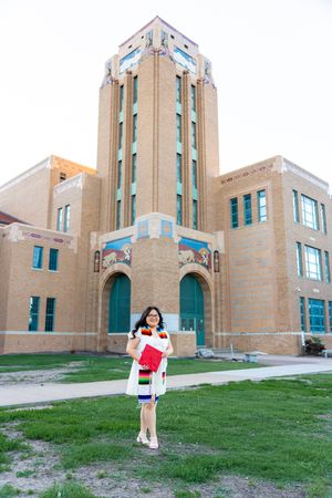 A graduate poses on a lawn in front of an Art Deco style building with a tall tower and turquoise accents.