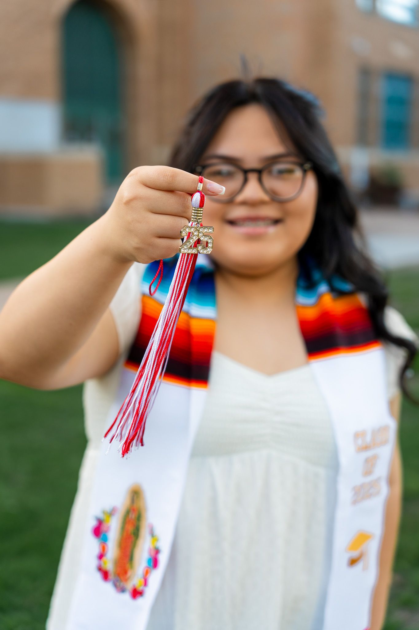 A graduate holds up a red and white tassel while wearing a colorful graduation stole and white gown.