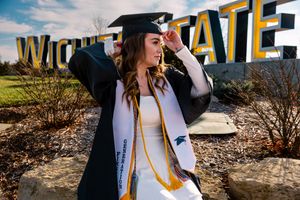 Graduate in white and gold regalia poses outdoors with diploma during commencement celebration.