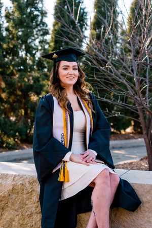 Graduate in cap and gown sits on stone wall during outdoor graduation photo session in natural setting.
