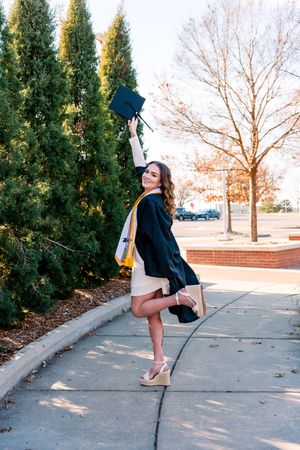 Graduate in cap and gown celebrates on campus sidewalk with trees and brick buildings in background on sunny day.