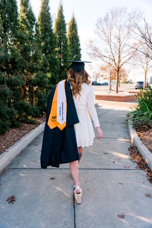 Graduate in cap and gown with gold honors sash walks on campus sidewalk wearing white dress and graduation attire.