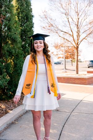 A graduate in a yellow stole and black cap poses outdoors near evergreen trees and bare branches at sunset.