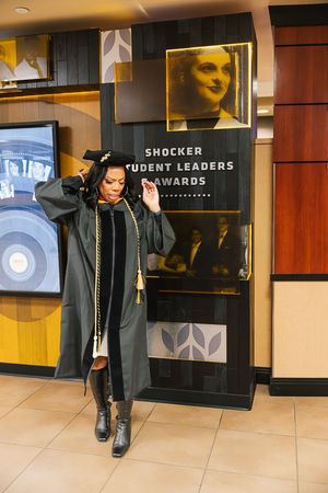 Graduate student in black cap and gown walks through hallway at university commencement ceremony.