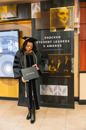 Graduate in black cap and gown walks past display wall showing student leaders and awards at university ceremony.