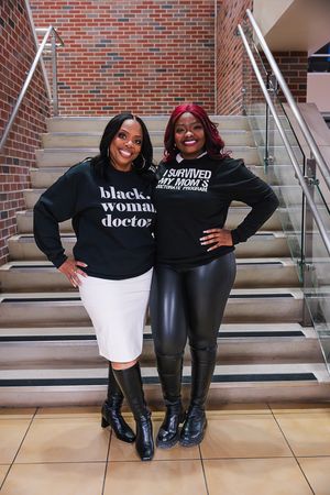 Two medical professionals in matching black shirts and boots sit together on stairs with a brick wall backdrop.