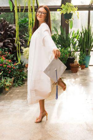 A person in a flowing white dress and high heels poses gracefully in a garden setting with potted plants and flowers.