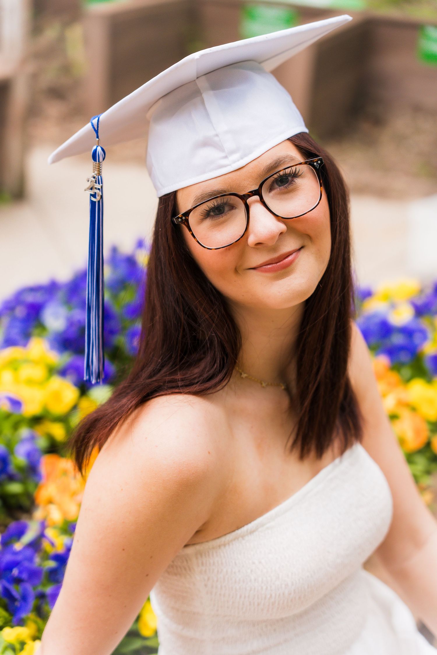 Graduate in white cap and gown poses near colorful spring flowers during outdoor graduation photo session.