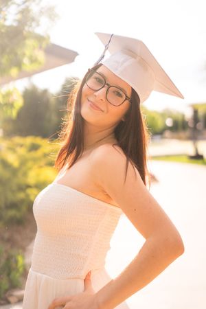 A graduate in a white dress and cap poses in warm sunlight during a golden hour photoshoot.